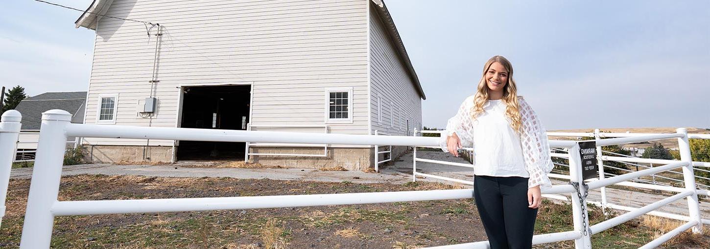 A woman stands next to a white fence with a white barn in the background.